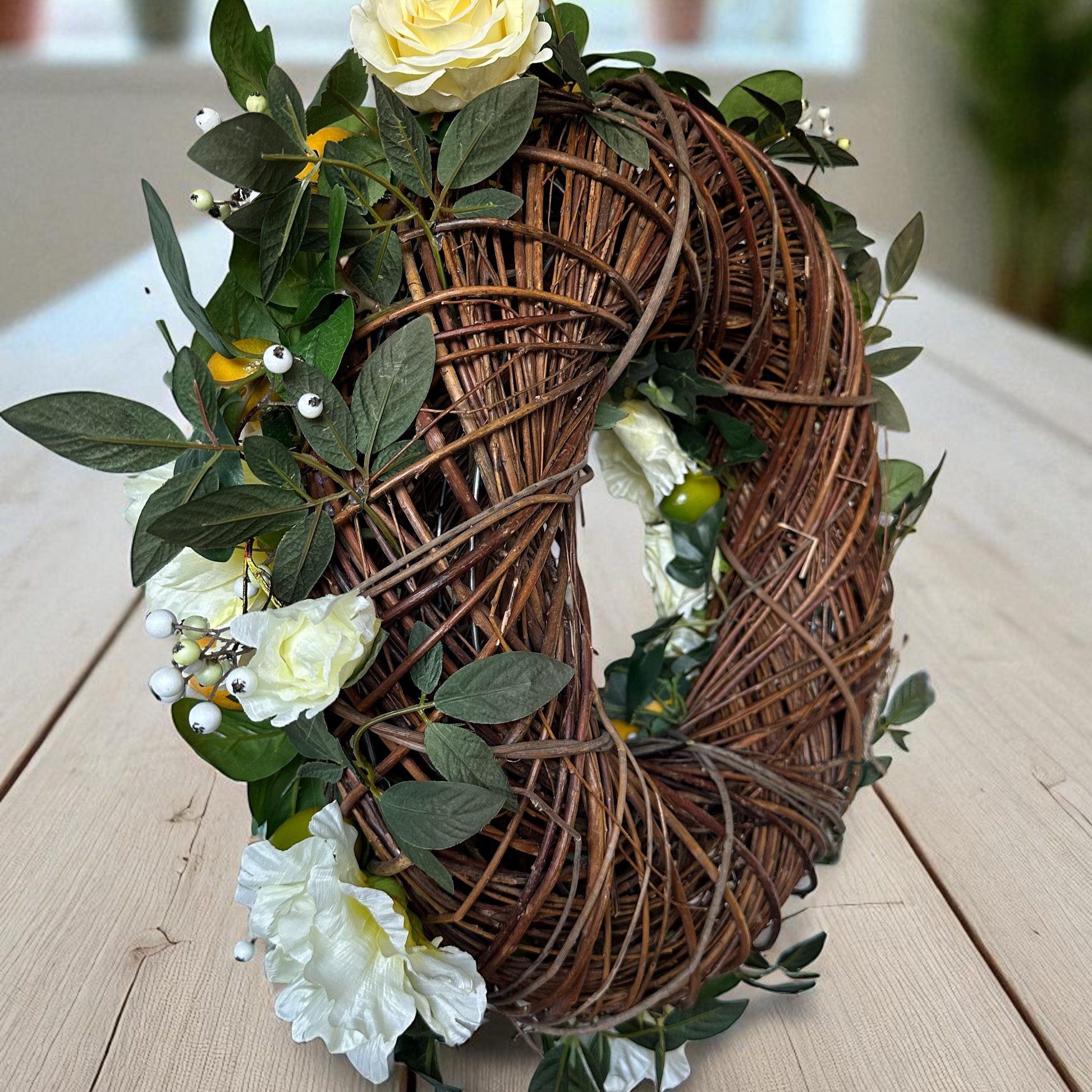 Een rustieke krans gemaakt van verweven bruine takken, versierd met witte rozen, groene bladeren en kleine witte bessen, geplaatst op een lichte houten tafel met een wazige binnenachtergrond