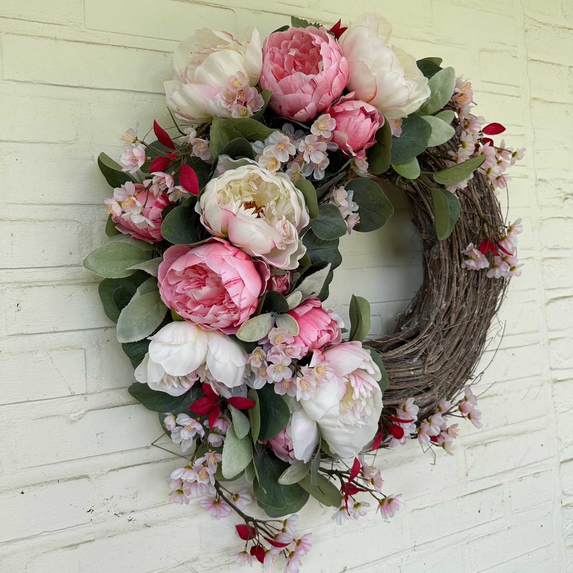 Handgemaakte luxe deurkrans met zijden pioenrozen in zachte blushroze en crèmekleurige tinten, gecombineerd met kunst kersenbloesems en fluweelachtig groen blad, zorgvuldig gerangschikt op een natuurlijke takkenbasis. Deze romantische, asymmetrische bloemenkrans van Jardin Enchanté straalt Franse elegantie en verfijnde charme uit. Perfect als luxe kunstbloemenkrans voor de voordeur, muurdecoratie of feestelijke gelegenheid, en ideaal voor liefhebbers van romantische, exclusieve seizoensdecoratie.