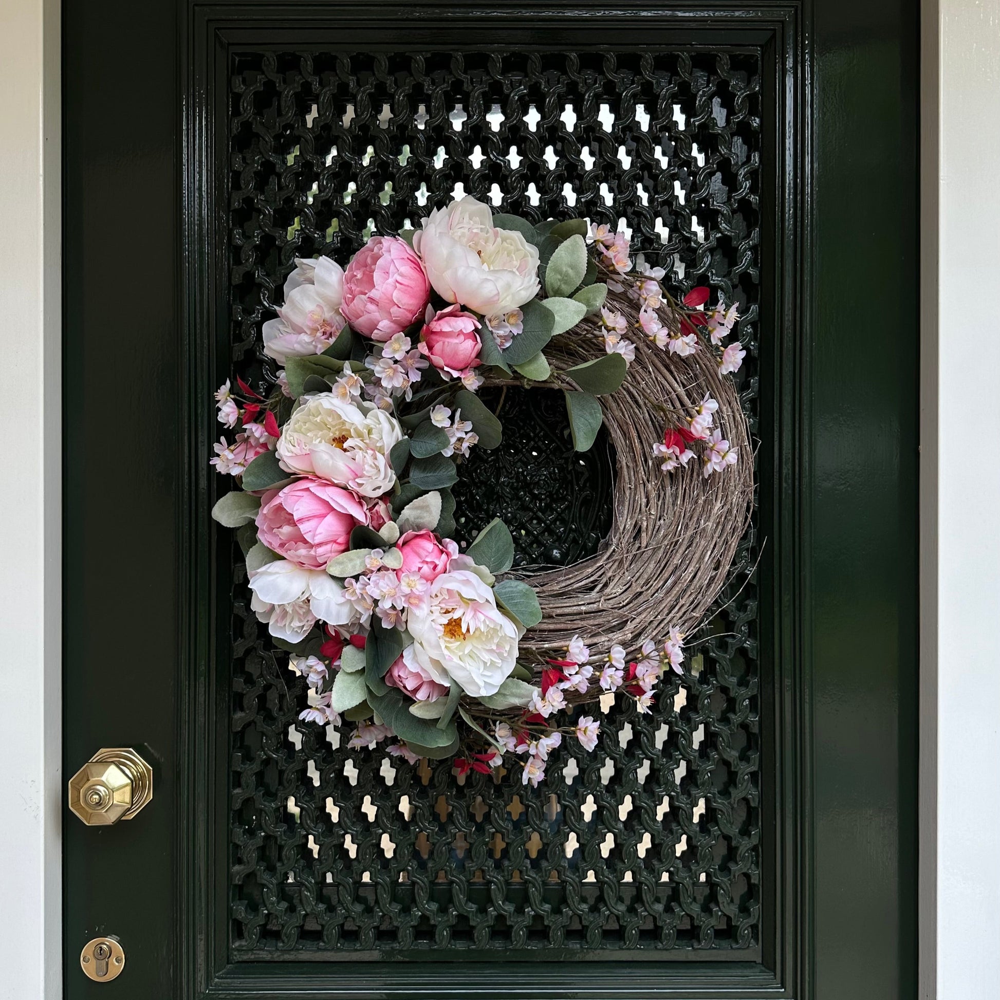 Handgemaakte luxe deurkrans met zijden pioenrozen in zachte blushroze en crèmekleurige tinten, gecombineerd met kunst kersenbloesems en fluweelachtig groen blad, zorgvuldig gerangschikt op een natuurlijke takkenbasis. Deze romantische, asymmetrische bloemenkrans van Jardin Enchanté straalt Franse elegantie en verfijnde charme uit. Perfect als luxe kunstbloemenkrans voor de voordeur, muurdecoratie of feestelijke gelegenheid, en ideaal voor liefhebbers van romantische, exclusieve seizoensdecoratie.