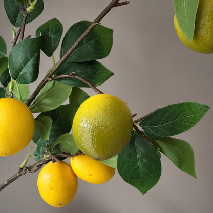 Detailed close-up of an artificial lemon branch featuring realistic yellow and green-yellow lemons with textured peels, deep green veined leaves and natural-looking brown twigs, photographed against a soft neutral background.