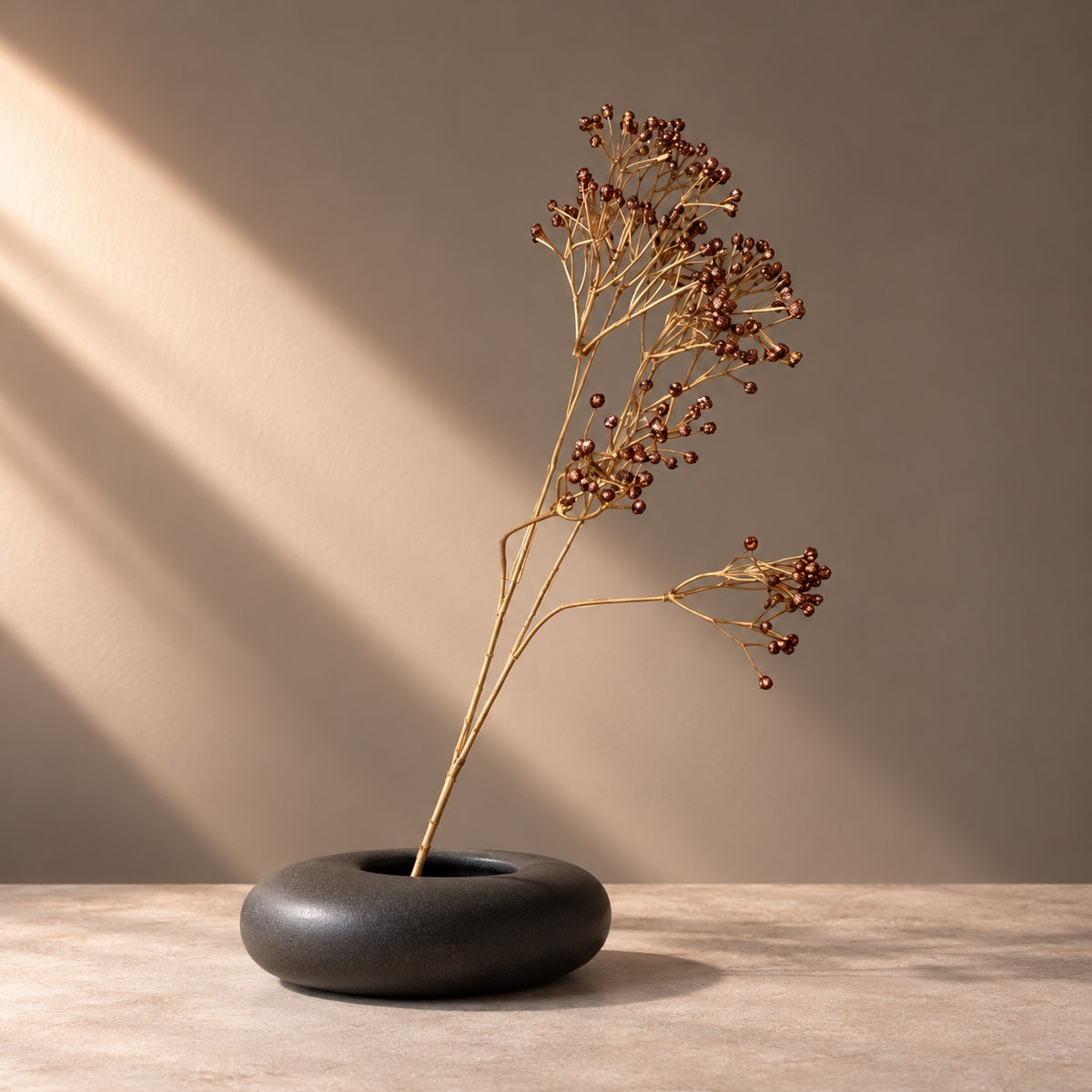 EN: Artificial copper berry stem with bronze berries styled in a low matte black sculptural vase. NL: Kunsttak met koperkleurige bessen in een lage matzwarte designvaas.