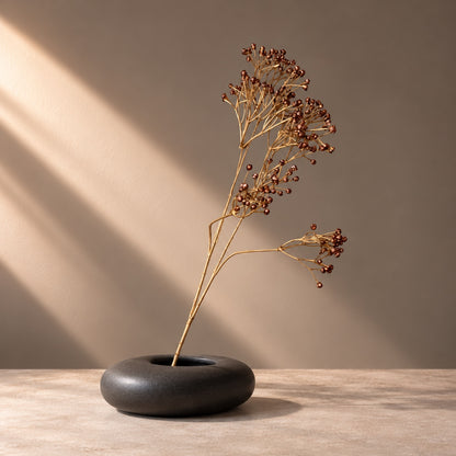 EN: Artificial copper berry stem with bronze berries styled in a low matte black sculptural vase. NL: Kunsttak met koperkleurige bessen in een lage matzwarte designvaas.