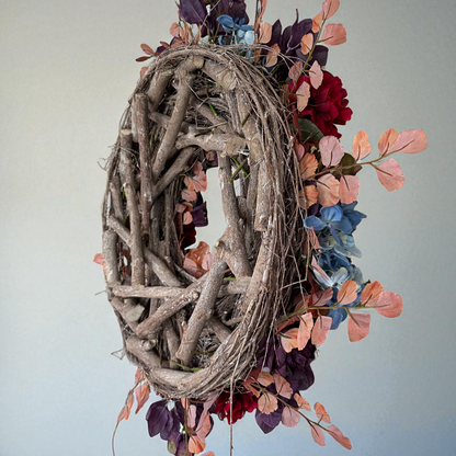 Side view of a handcrafted autumn wreath with a rustic grapevine twig base, decorated with burgundy peonies, blue hydrangeas, peach blossoms, and autumn foliage. The wreath showcases its natural wooden structure, highlighting the artisanal quality and seasonal charm of this fall door decoration.