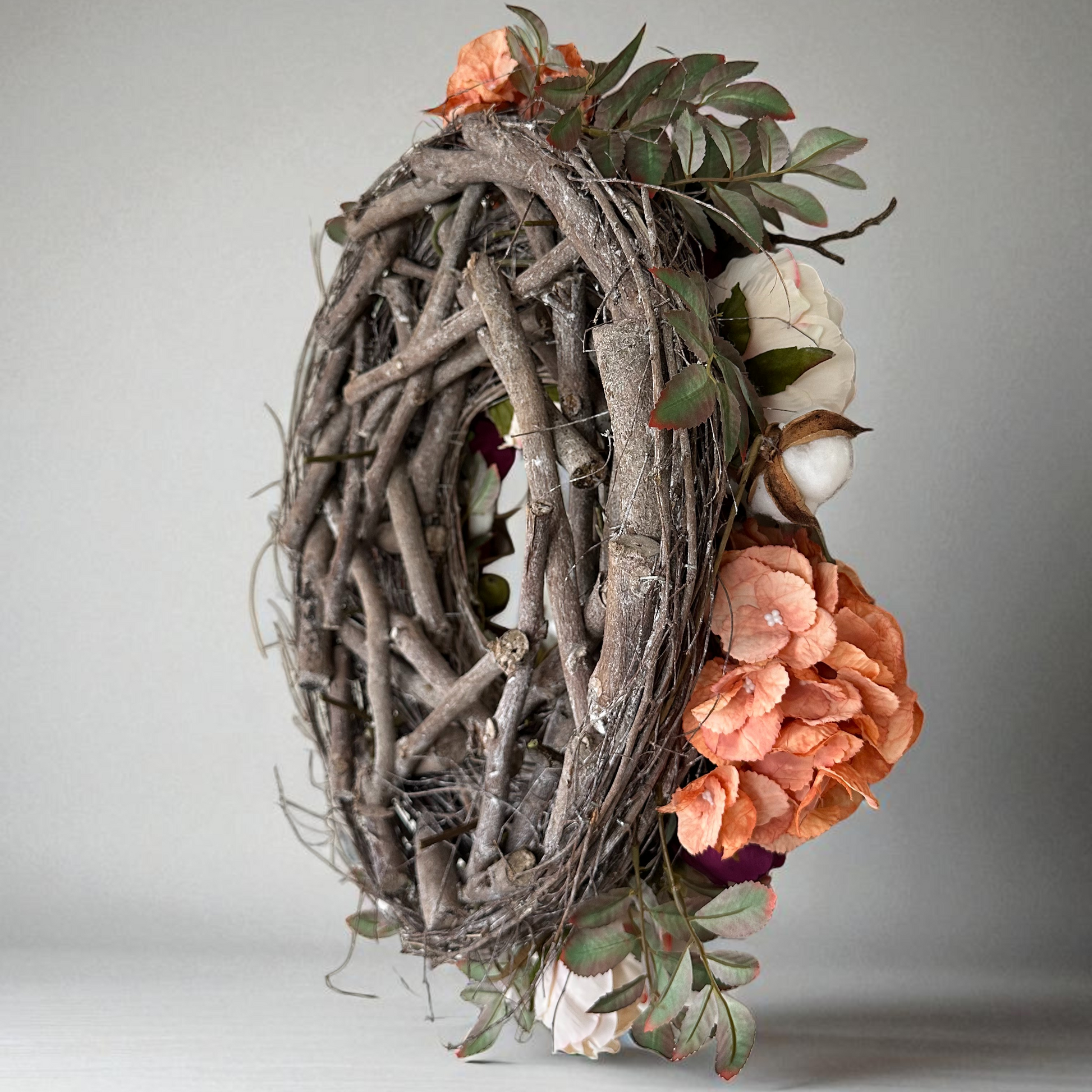 Side view of a handmade autumn wreath with rustic grapevine twig base, decorated with peach hydrangeas, burgundy peonies, blush roses, green apple accents, and soft cotton flowers. Elegant fall floral door wreath for farmhouse decor, front door styling, and seasonal home decoration.