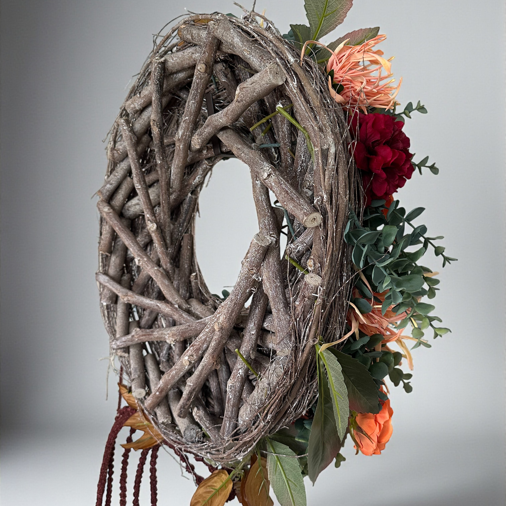 Side view of the Falling Roses Wreath with rustic twig base, decorated with orange and burgundy roses, eucalyptus leaves, and wilted spider mum flowers, highlighting the natural handcrafted design.