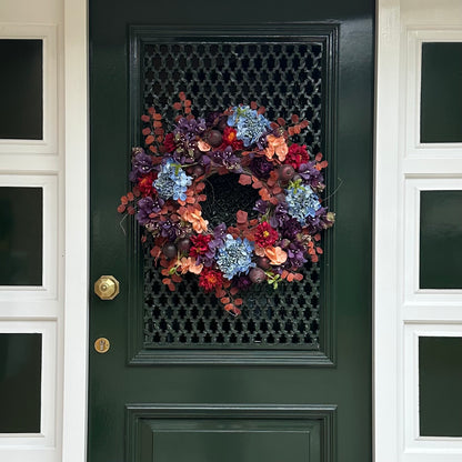 Handgemaakte herfstkrans aan voordeur met blauwe hortensia’s, bordeauxrode pioenrozen, perzikbloemen en rode zinnia’s, aangevuld met sierappels en herfstbladeren. Een elegante herfstdecoratie voor de voordeur die warmte en stijl toevoegt aan elk huis.