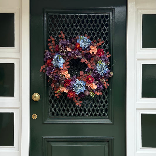 Handgemaakte herfstkrans aan voordeur met blauwe hortensia’s, bordeauxrode pioenrozen, perzikbloemen en rode zinnia’s, aangevuld met sierappels en herfstbladeren. Een elegante herfstdecoratie voor de voordeur die warmte en stijl toevoegt aan elk huis.