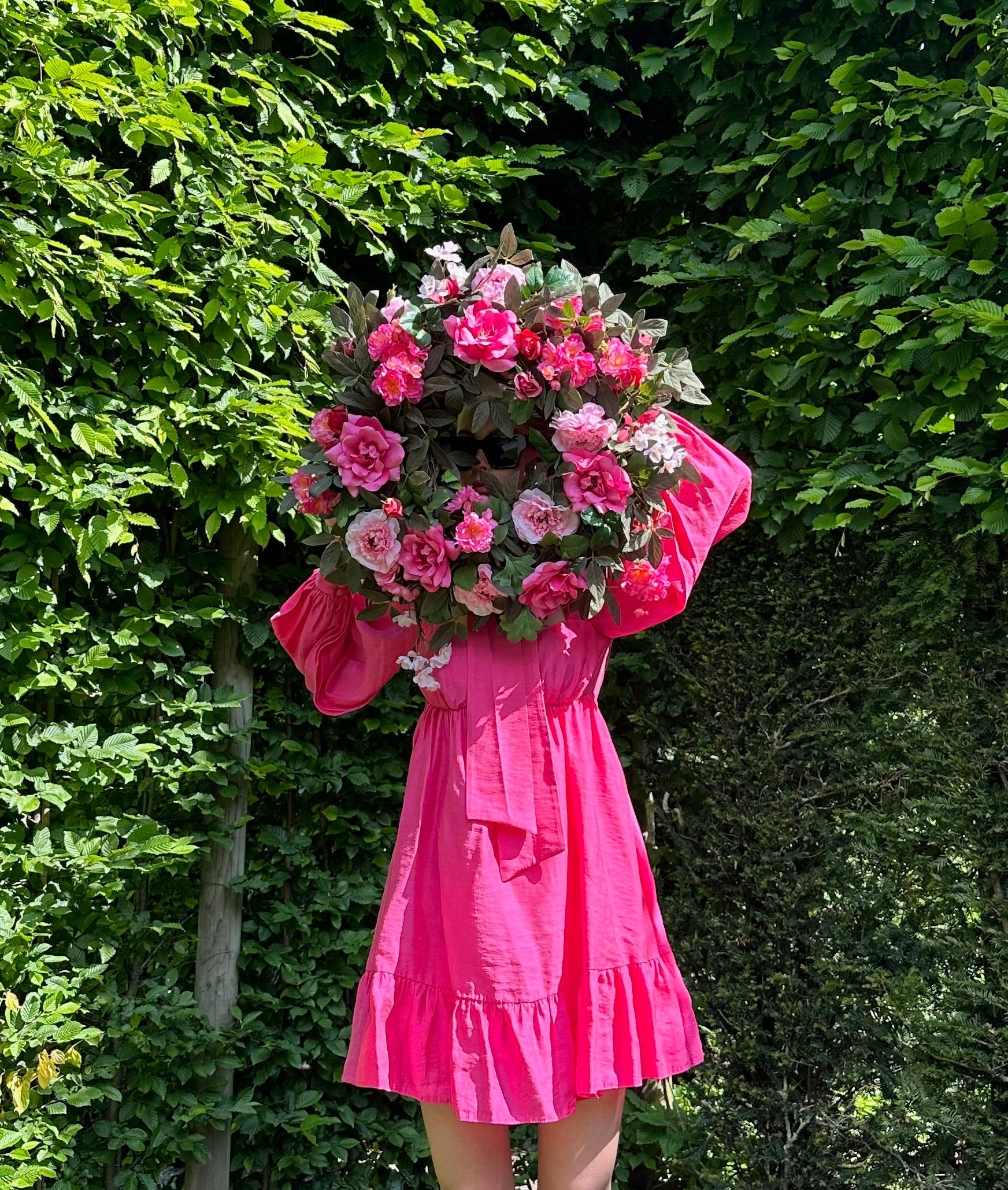 Person in a pink dress holding a large door wreath made of high quality faux flowers against a green hedge.