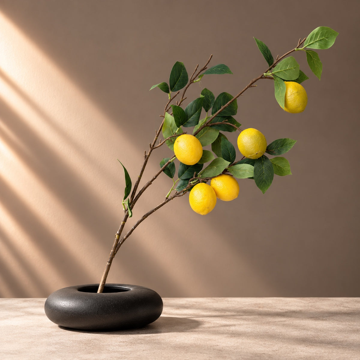EN: Single 85 cm artificial lemon branch with five realistic yellow lemons, detailed green leaves and slender brown twigs, styled diagonally in a low round black vase on a neutral stone surface with warm diagonal sunlight. NL: Enkele kunstcitroentak van 85 cm met vijf levensechte gele citroenen, gedetailleerde groene bladeren en slanke bruine twijgen, schuin gestyled in een lage ronde zwarte vaas op een neutraal stenen oppervlak met schuin invallend zonlicht.