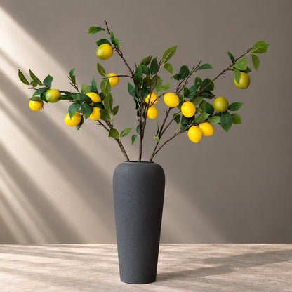 EN: Artificial lemon branches arranged in a tall matte charcoal vase, with vivid yellow lemons and rich green leaves against a soft taupe wall and stone tabletop in natural sunlight. NL: Kunsttakken met citroenen in een hoge mat antracietkleurige vaas, met heldergele citroenen en diepgroene bladeren tegen een zachte taupekleurige muur en stenen tafelblad in natuurlijk zonlicht.