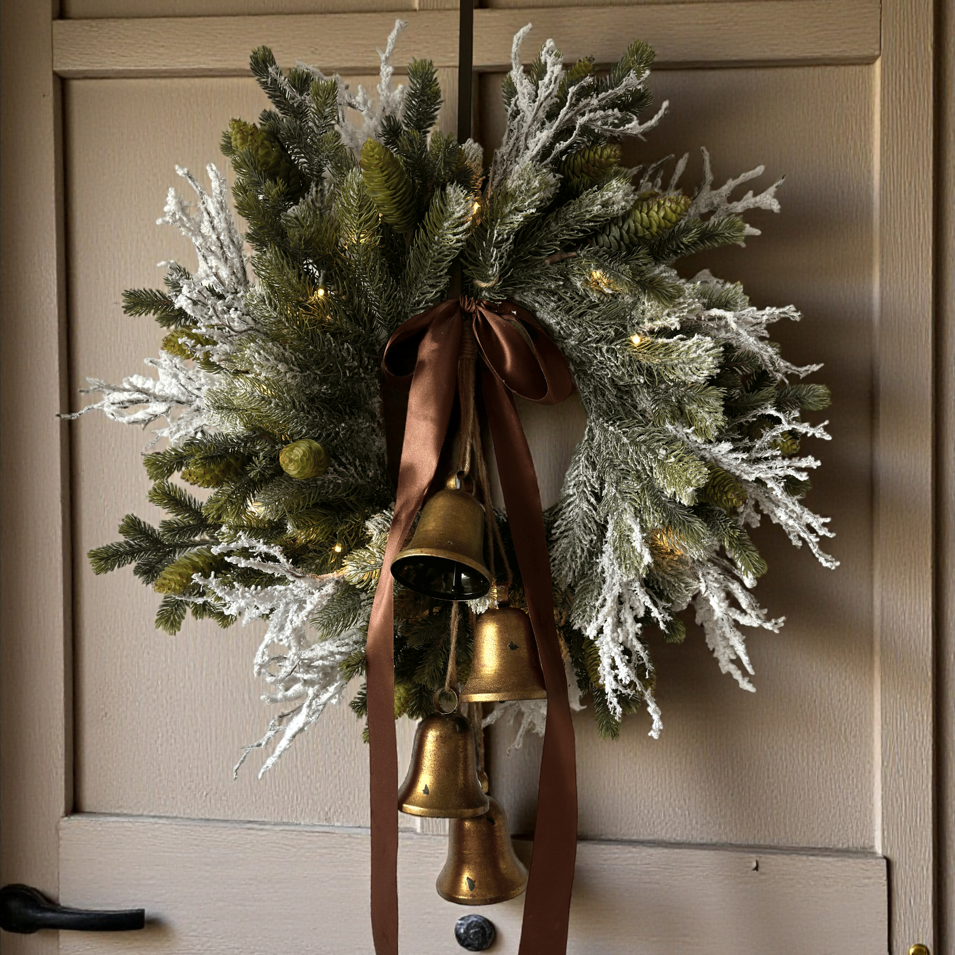 Stijlvolle kunstkerstkrans met gouden belletjes, bruine satijnen strik en warme LED-verlichting, opgehangen aan een moderne grijze voordeur – luxe winterdecoratie voor buiten.