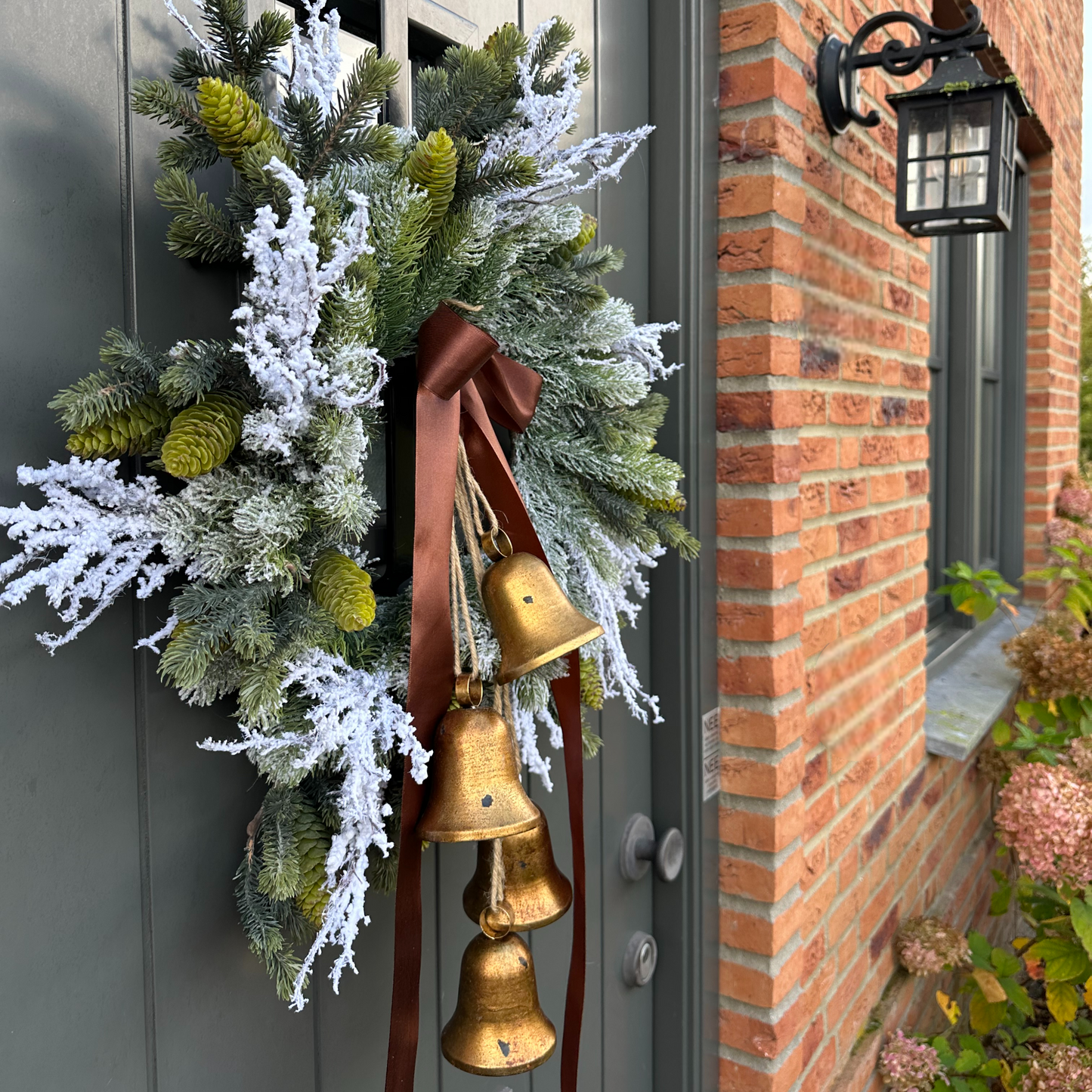 Stijlvolle kunstkerstkrans met gouden belletjes, bruine satijnen strik en warme LED-verlichting, opgehangen aan een moderne grijze voordeur – luxe winterdecoratie voor buiten.