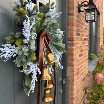 Stijlvolle kunstkerstkrans met gouden belletjes, bruine satijnen strik en warme LED-verlichting, opgehangen aan een moderne grijze voordeur – luxe winterdecoratie voor buiten.