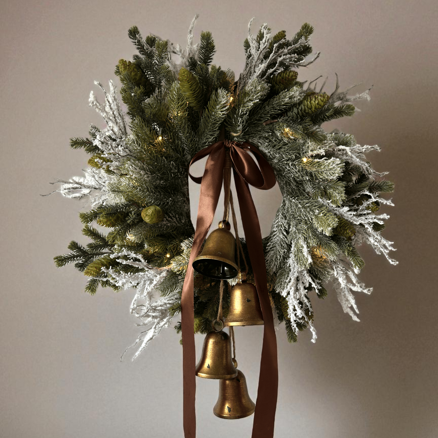 Stijlvolle kunstkerstkrans met gouden belletjes, bruine satijnen strik en warme LED-verlichting, opgehangen aan een moderne grijze voordeur – luxe winterdecoratie voor buiten.