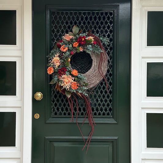 Handgemaakte herfstkrans Falling Roses op een donkere voordeur, versierd met oranje rozen, dieprode bloemen, eucalyptus en verwelkte spinnenchrysanten. De natuurlijke takkenbasis en lange sierlijke hangende elementen geven deze luxe deurkrans een elegante en seizoensgebonden uitstraling, ideaal voor herfstdecoratie.