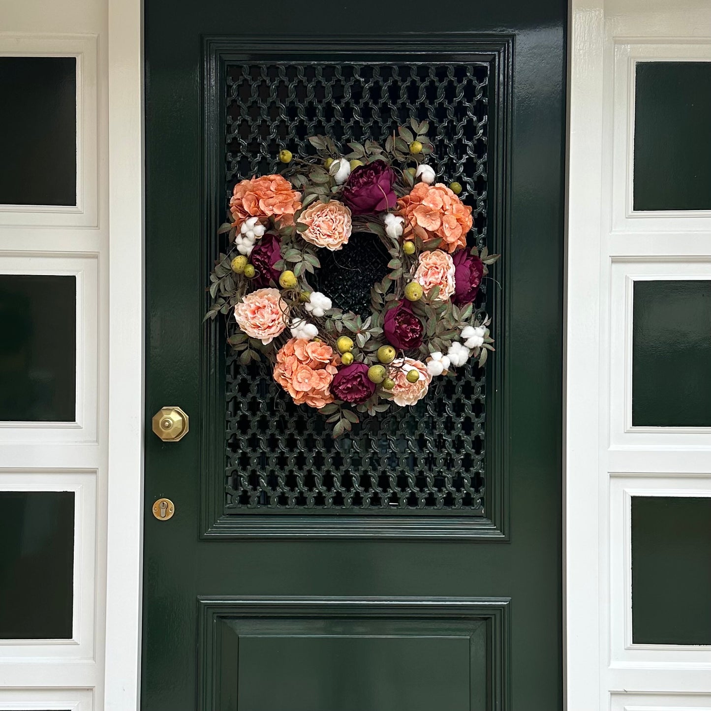 Voordeur versierd met een luxe herfstkrans van perzik hortensia’s, bordeaux pioenen, blush rozen, groene appeltjes en witte katoenbloemen op een basis van druivenranken. Elegante herfstkrans voor de voordeur, seizoensgebonden woondecoratie, landelijke stijl en feestelijke entree decoratie.
