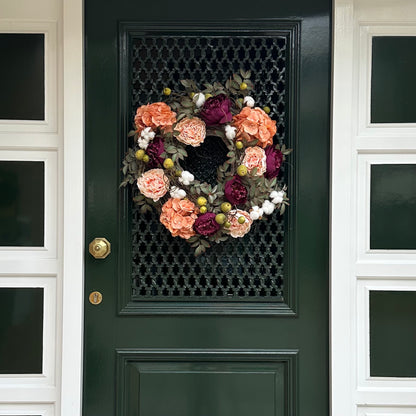 Voordeur versierd met een luxe herfstkrans van perzik hortensia’s, bordeaux pioenen, blush rozen, groene appeltjes en witte katoenbloemen op een basis van druivenranken. Elegante herfstkrans voor de voordeur, seizoensgebonden woondecoratie, landelijke stijl en feestelijke entree decoratie.