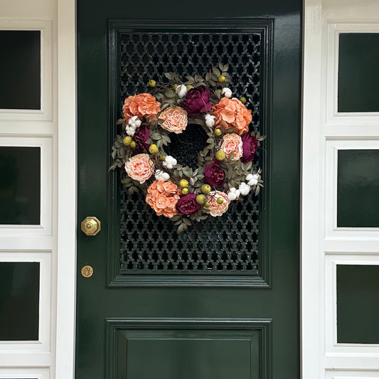Voordeur versierd met een luxe herfstkrans van perzik hortensia’s, bordeaux pioenen, blush rozen, groene appeltjes en witte katoenbloemen op een basis van druivenranken. Elegante herfstkrans voor de voordeur, seizoensgebonden woondecoratie, landelijke stijl en feestelijke entree decoratie.
