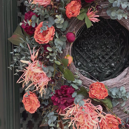 Handgemaakte herfstkrans Falling Roses op een donkere voordeur, versierd met oranje rozen, dieprode bloemen, eucalyptus en verwelkte spinnenchrysanten. De natuurlijke takkenbasis en lange sierlijke hangende elementen geven deze luxe deurkrans een elegante en seizoensgebonden uitstraling, ideaal voor herfstdecoratie.
