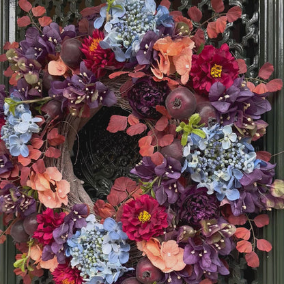 Handgemaakte herfstkrans aan voordeur met blauwe hortensia’s, bordeauxrode pioenrozen, perzikbloemen en rode zinnia’s, aangevuld met sierappels en herfstbladeren. Een elegante herfstdecoratie voor de voordeur die warmte en stijl toevoegt aan elk huis.