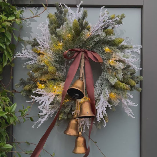 Stijlvolle kunstkerstkrans met gouden belletjes, bruine satijnen strik en warme LED-verlichting, opgehangen aan een moderne grijze voordeur – luxe winterdecoratie voor buiten