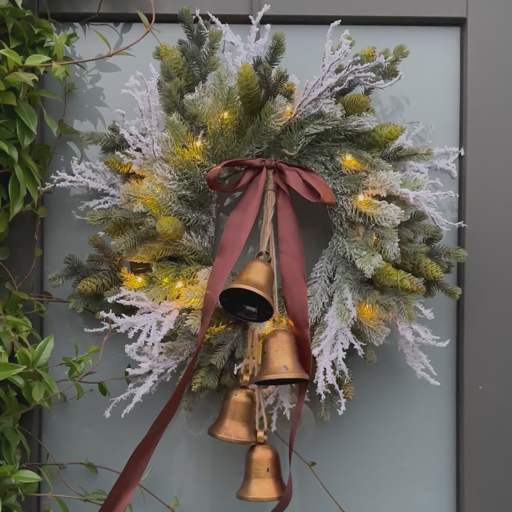Stijlvolle kunstkerstkrans met gouden belletjes, bruine satijnen strik en warme LED-verlichting, opgehangen aan een moderne grijze voordeur – luxe winterdecoratie voor buiten