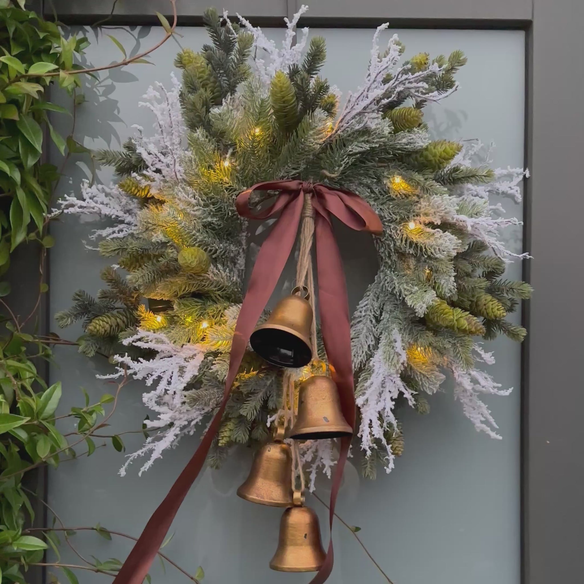 Stijlvolle kunstkerstkrans met gouden belletjes, bruine satijnen strik en warme LED-verlichting, opgehangen aan een moderne grijze voordeur – luxe winterdecoratie voor buiten
