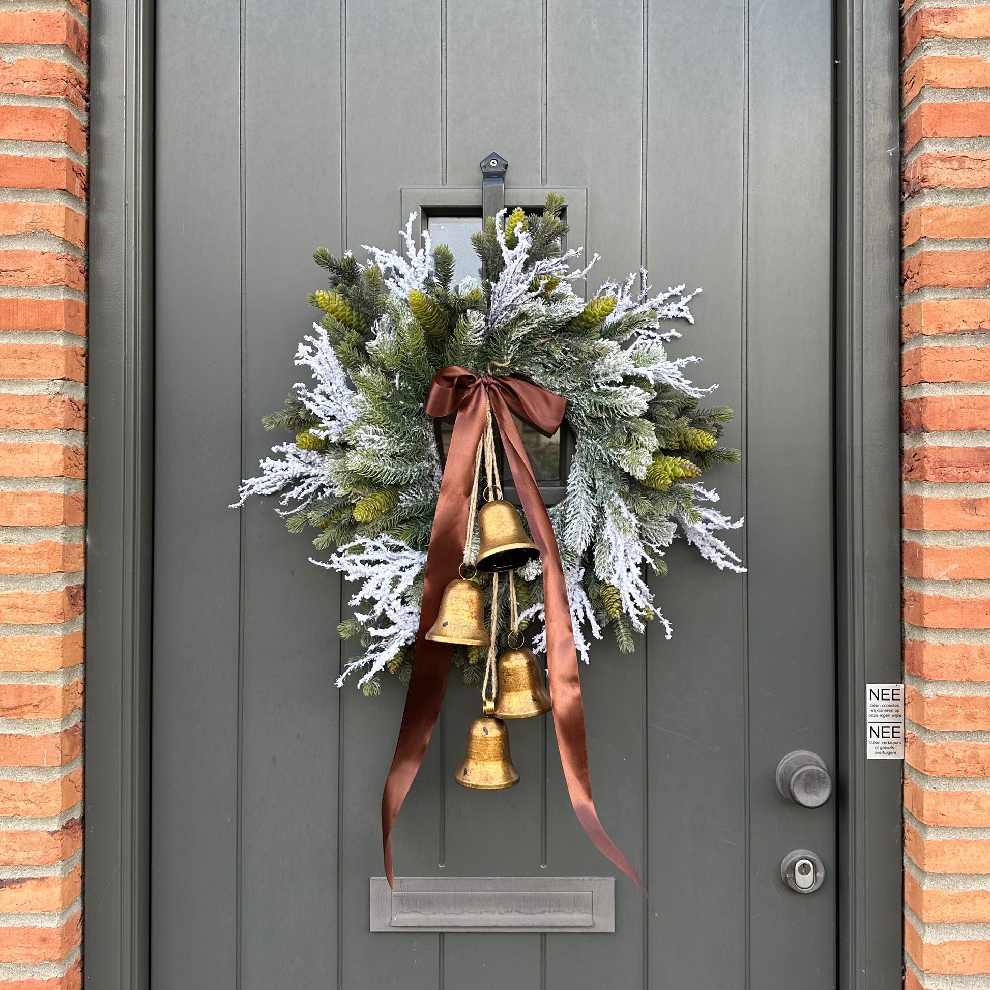Stijlvolle kunstkerstkrans met gouden belletjes en bruine satijnen strik, opgehangen aan een moderne grijze voordeur – luxe winterdecoratie voor buiten
